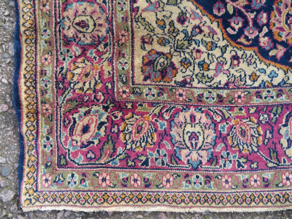 A rectangular hand-knotted wool rug featuring a central indigo floral medallion, cream spandrels, and a magenta border. View III