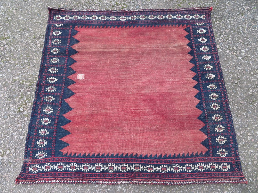 A square antique Afshar flatweave sofreh rug with a faded red central field and serrated indigo blue geometric borders.