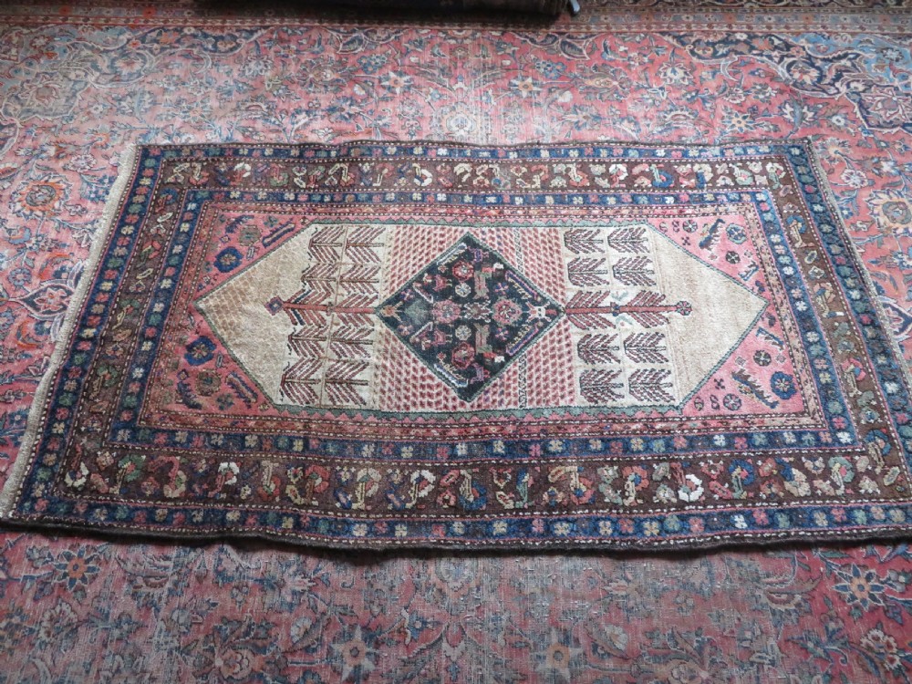 An antique Kurdish tribal rug featuring a cream field with stylized tree motifs, a central navy diamond medallion, and terracotta pink geometric borders. View XVII