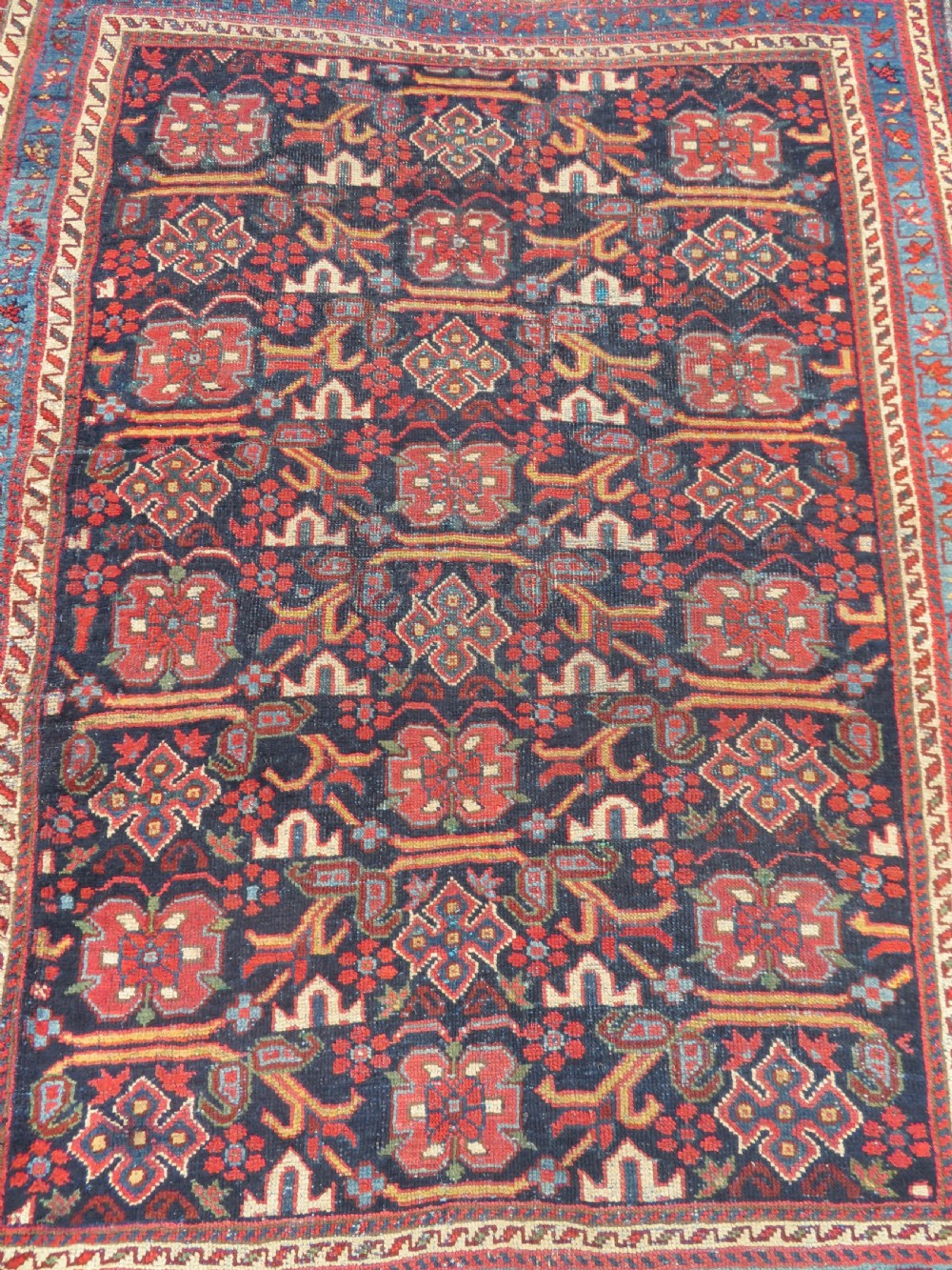 A rectangular hand-knotted antique Afshar rug featuring geometric floral motifs in terracotta red and sky blue on a deep navy blue background. View III
