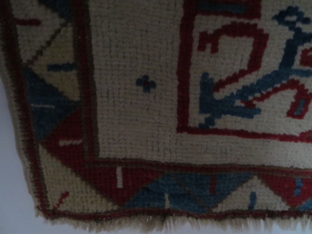 An antique Anatolian Konya wool rug featuring geometric red and blue symbols on a cream field with a sawtooth border. View X