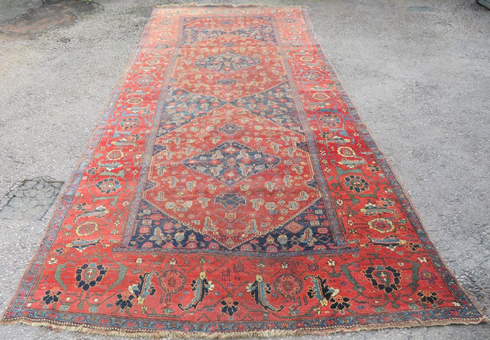 An antique Kurdish Bidjar Kelleh hand-knotted carpet featuring geometric diamond medallions on a terracotta red field with dark navy blue borders.