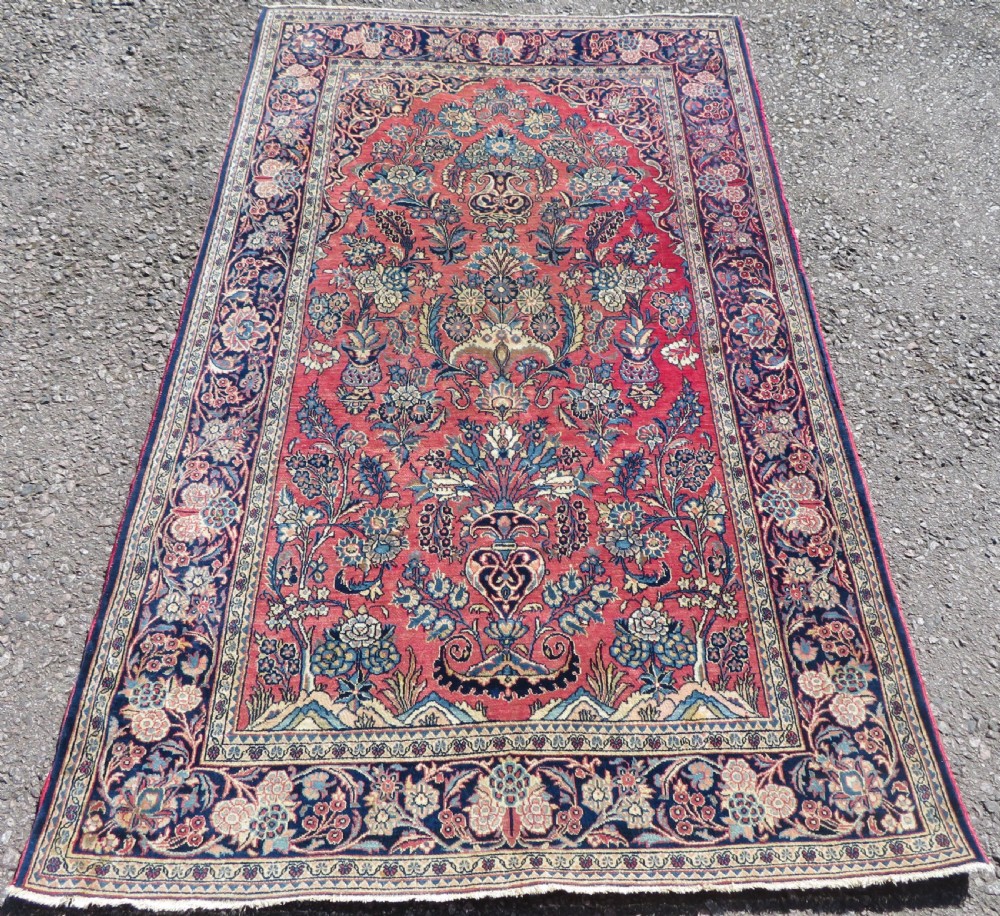 A rectangular hand-knotted Persian Kashan rug featuring a detailed crimson red field adorned with floral and vase motifs surrounded by a deep navy blue ornate border.