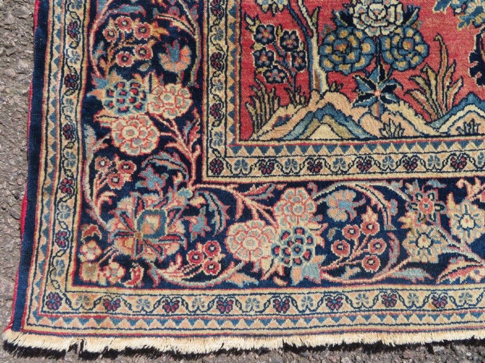 A rectangular hand-knotted Persian Kashan rug featuring a detailed crimson red field adorned with floral and vase motifs surrounded by a deep navy blue ornate border. View VI