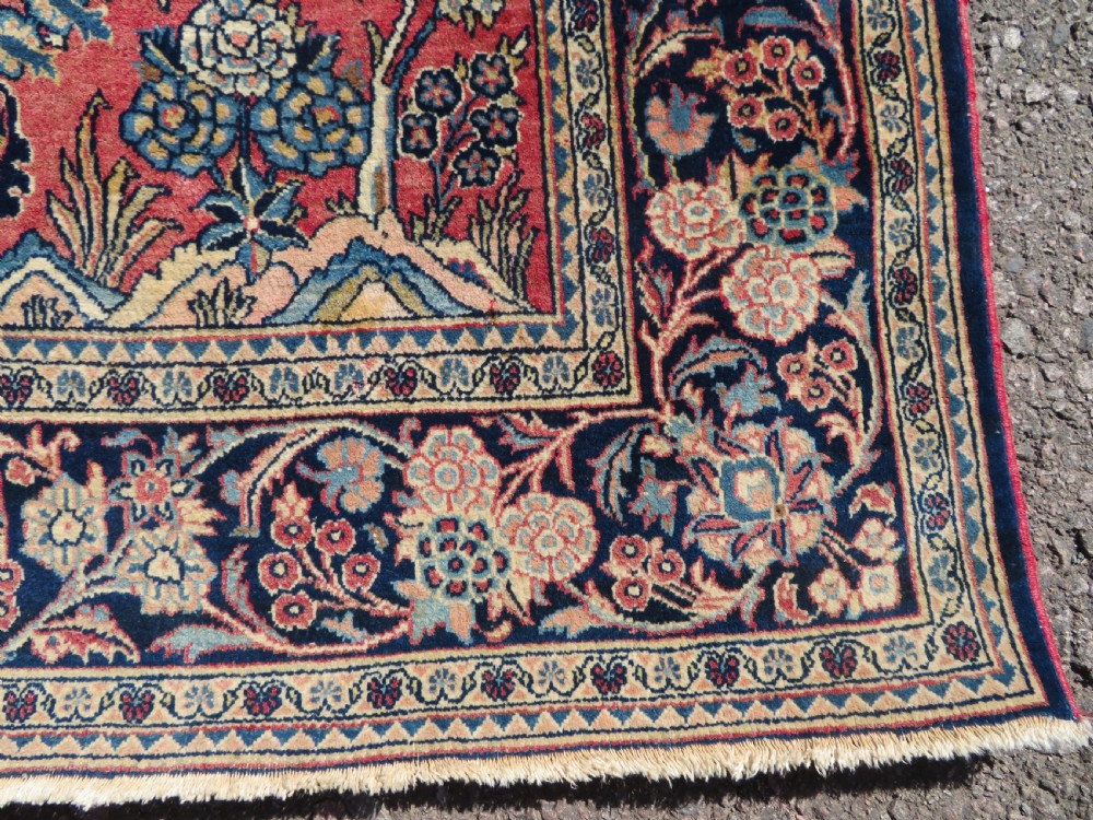 A rectangular hand-knotted Persian Kashan rug featuring a detailed crimson red field adorned with floral and vase motifs surrounded by a deep navy blue ornate border. View VII