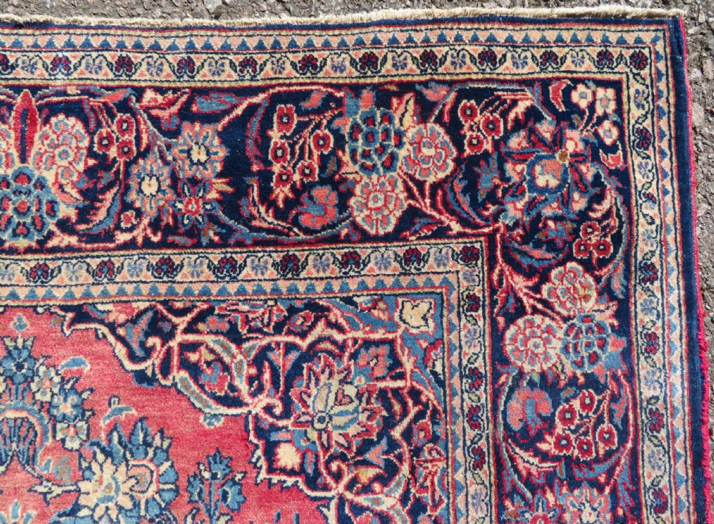 A rectangular hand-knotted Persian Kashan rug featuring a detailed crimson red field adorned with floral and vase motifs surrounded by a deep navy blue ornate border. View VIII