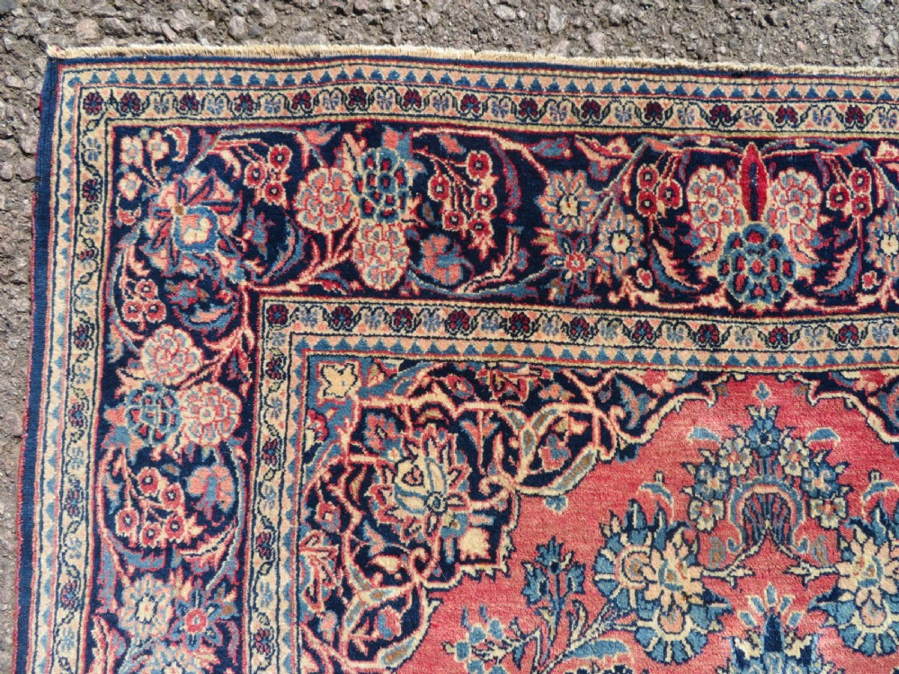 A rectangular hand-knotted Persian Kashan rug featuring a detailed crimson red field adorned with floral and vase motifs surrounded by a deep navy blue ornate border. View IX