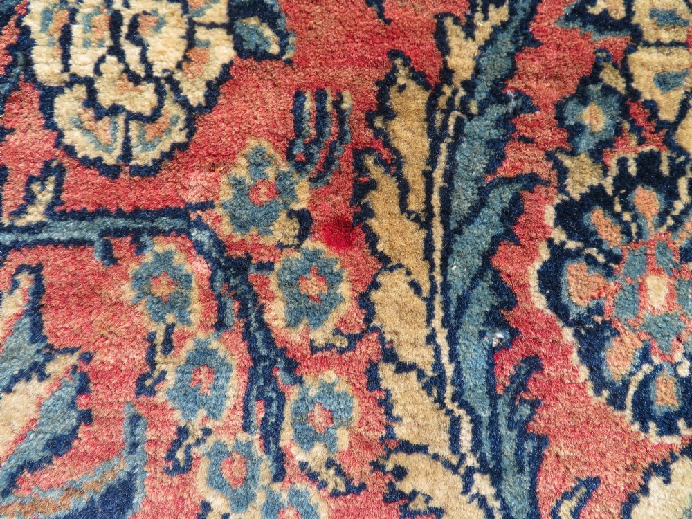 A rectangular hand-knotted Persian Kashan rug featuring a detailed crimson red field adorned with floral and vase motifs surrounded by a deep navy blue ornate border. View X