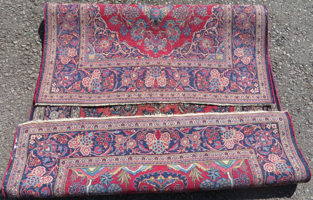 A rectangular hand-knotted Persian Kashan rug featuring a detailed crimson red field adorned with floral and vase motifs surrounded by a deep navy blue ornate border. View XII