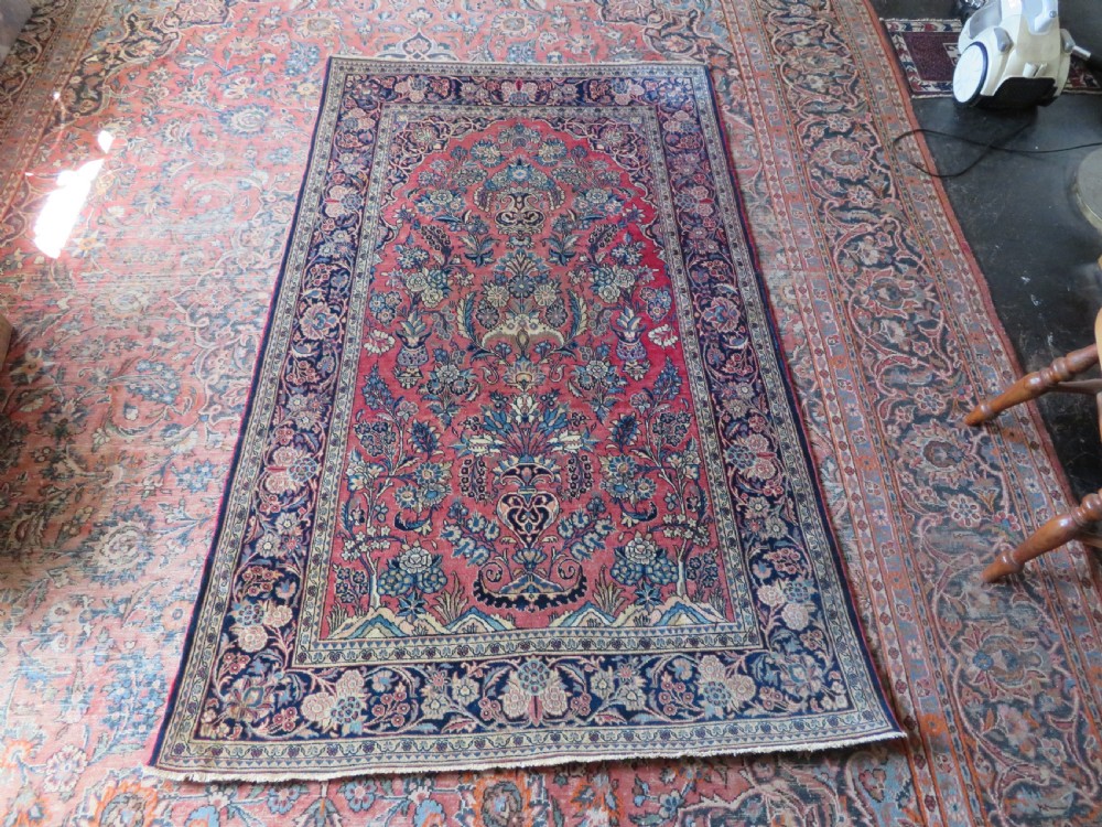 A rectangular hand-knotted Persian Kashan rug featuring a detailed crimson red field adorned with floral and vase motifs surrounded by a deep navy blue ornate border. View XVI