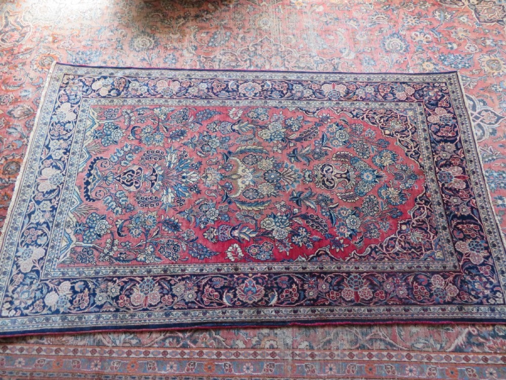 A rectangular hand-knotted Persian Kashan rug featuring a detailed crimson red field adorned with floral and vase motifs surrounded by a deep navy blue ornate border. View XVII