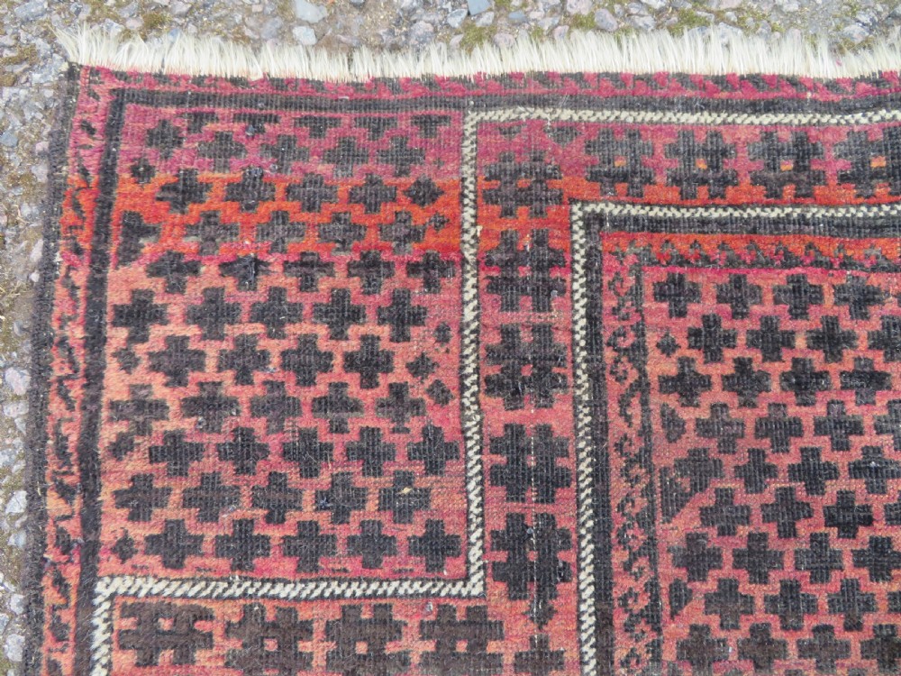 An antique Baluch prayer rug featuring a dark red and brown geometric grid pattern inside a defined mihrab arch with woven fringed ends. View III