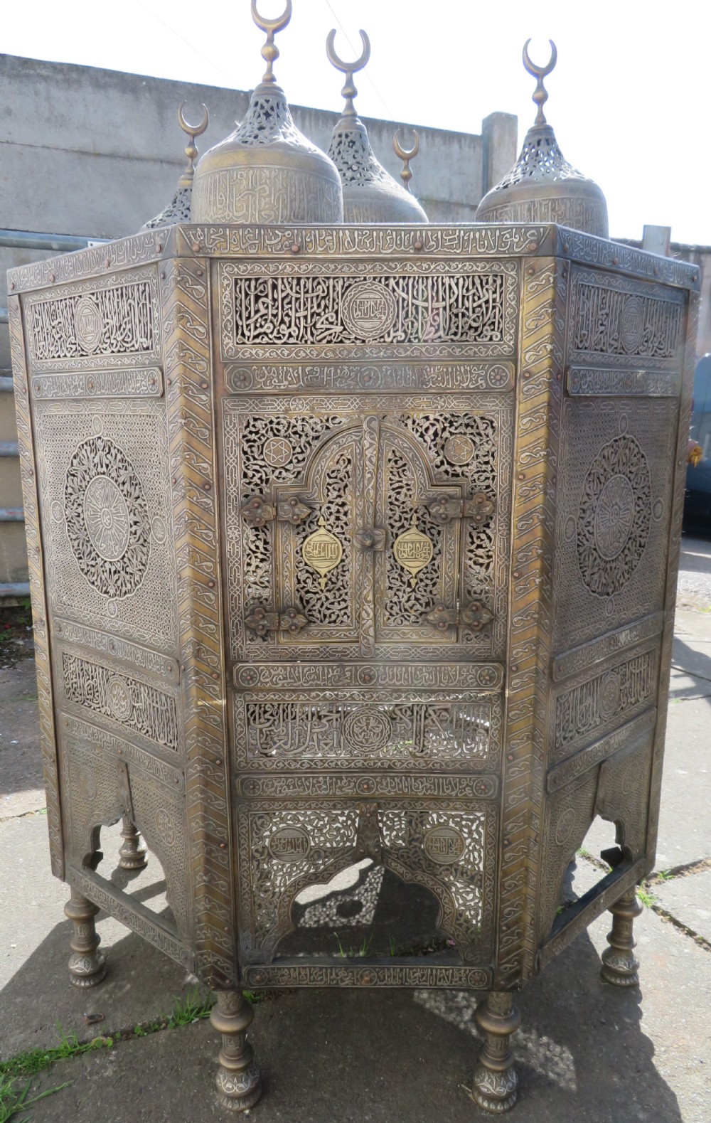 An octagonal brass and copper Cairoware Quran stand featuring Arabic calligraphy, pierced fretwork panels, silver inlay, and top dome finials crowned with crescent moons.