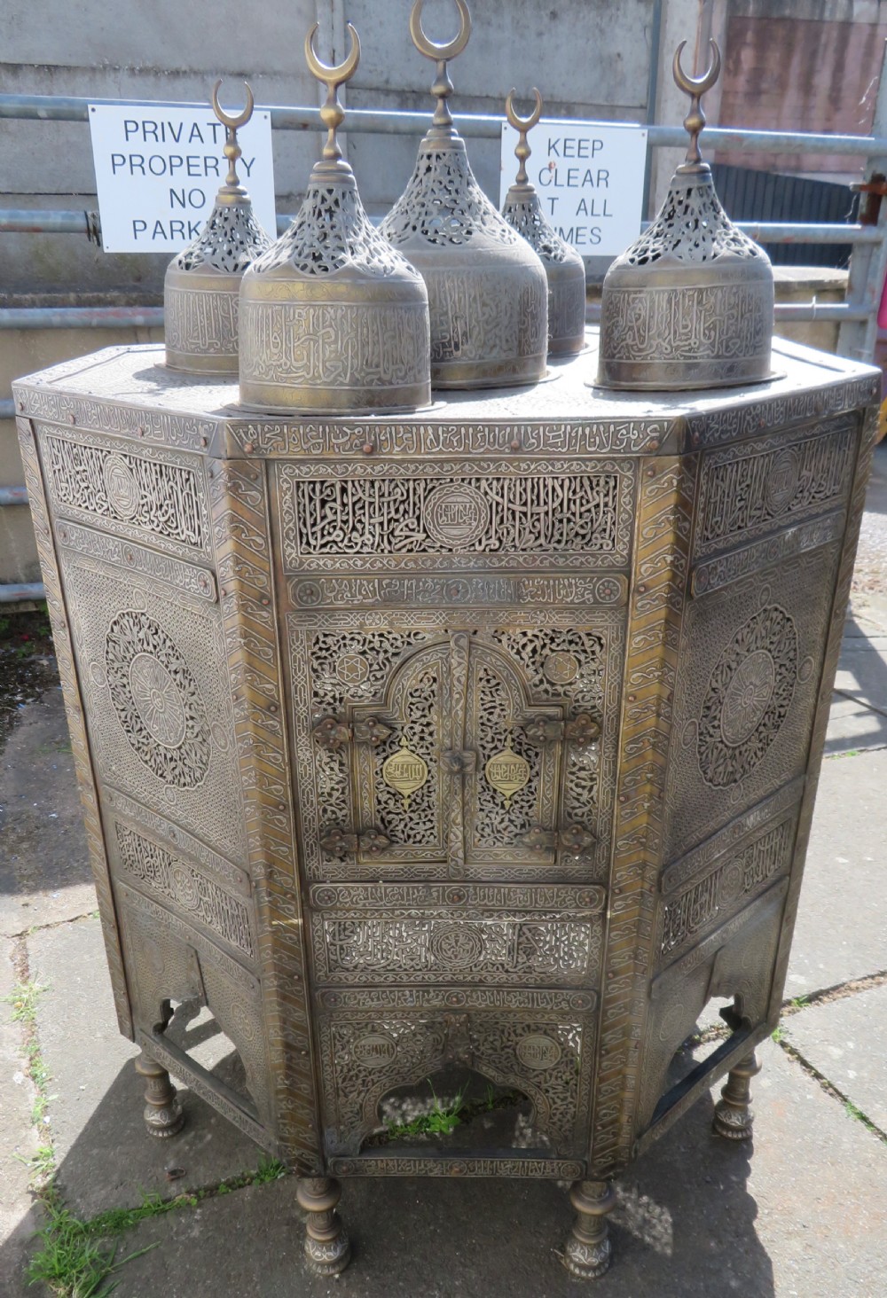 An octagonal brass and copper Cairoware Quran stand featuring Arabic calligraphy, pierced fretwork panels, silver inlay, and top dome finials crowned with crescent moons. View II