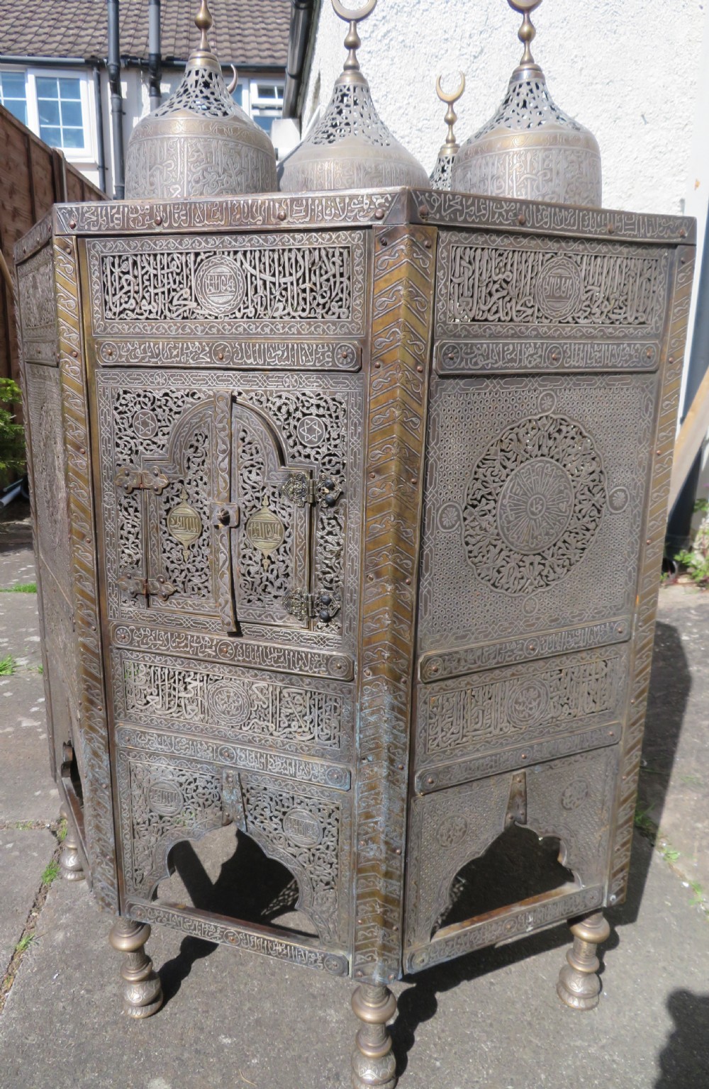 An octagonal brass and copper Cairoware Quran stand featuring Arabic calligraphy, pierced fretwork panels, silver inlay, and top dome finials crowned with crescent moons. View V