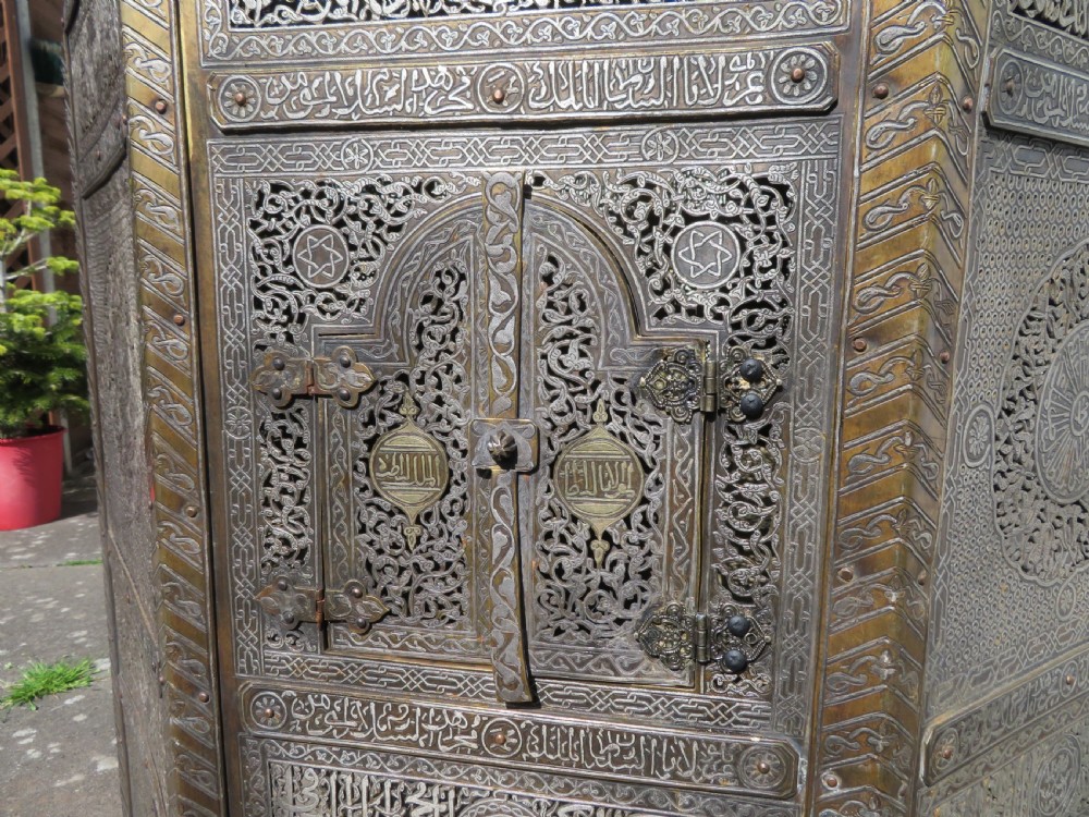 An octagonal brass and copper Cairoware Quran stand featuring Arabic calligraphy, pierced fretwork panels, silver inlay, and top dome finials crowned with crescent moons. View VI