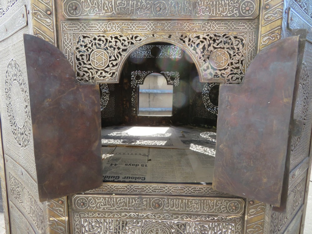 An octagonal brass and copper Cairoware Quran stand featuring Arabic calligraphy, pierced fretwork panels, silver inlay, and top dome finials crowned with crescent moons. View VIII