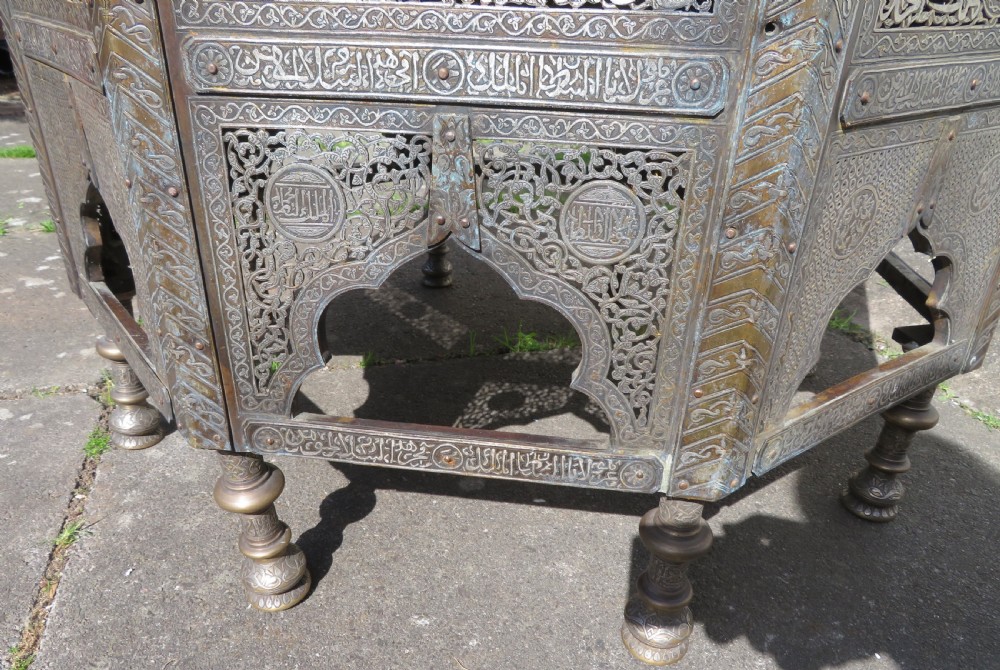 An octagonal brass and copper Cairoware Quran stand featuring Arabic calligraphy, pierced fretwork panels, silver inlay, and top dome finials crowned with crescent moons. View IX
