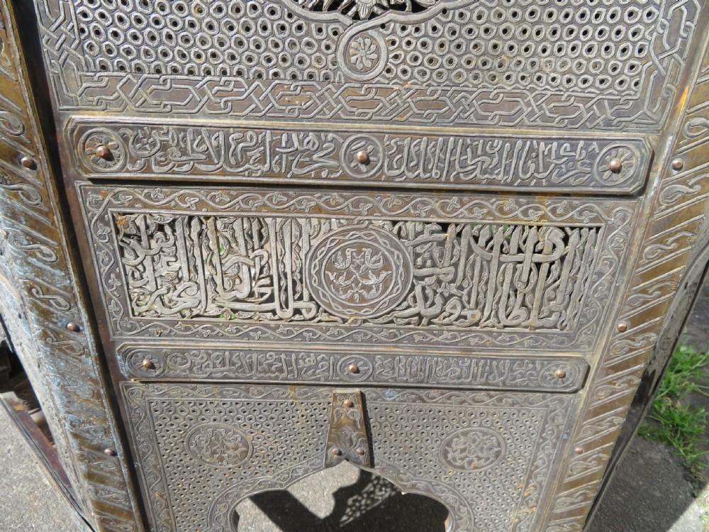 An octagonal brass and copper Cairoware Quran stand featuring Arabic calligraphy, pierced fretwork panels, silver inlay, and top dome finials crowned with crescent moons. View X