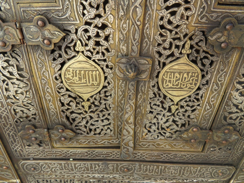An octagonal brass and copper Cairoware Quran stand featuring Arabic calligraphy, pierced fretwork panels, silver inlay, and top dome finials crowned with crescent moons. View XVI