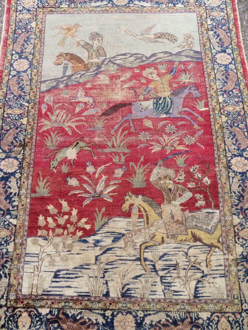 A 19th-century West Anatolian silk rug featuring a red central panel depicting horseback hunters, surrounded by a floral and animal motif navy border. View II