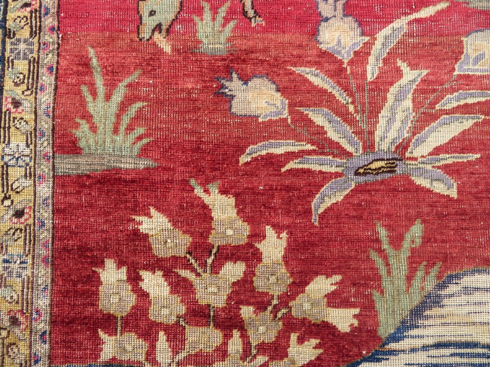 A 19th-century West Anatolian silk rug featuring a red central panel depicting horseback hunters, surrounded by a floral and animal motif navy border. View IV
