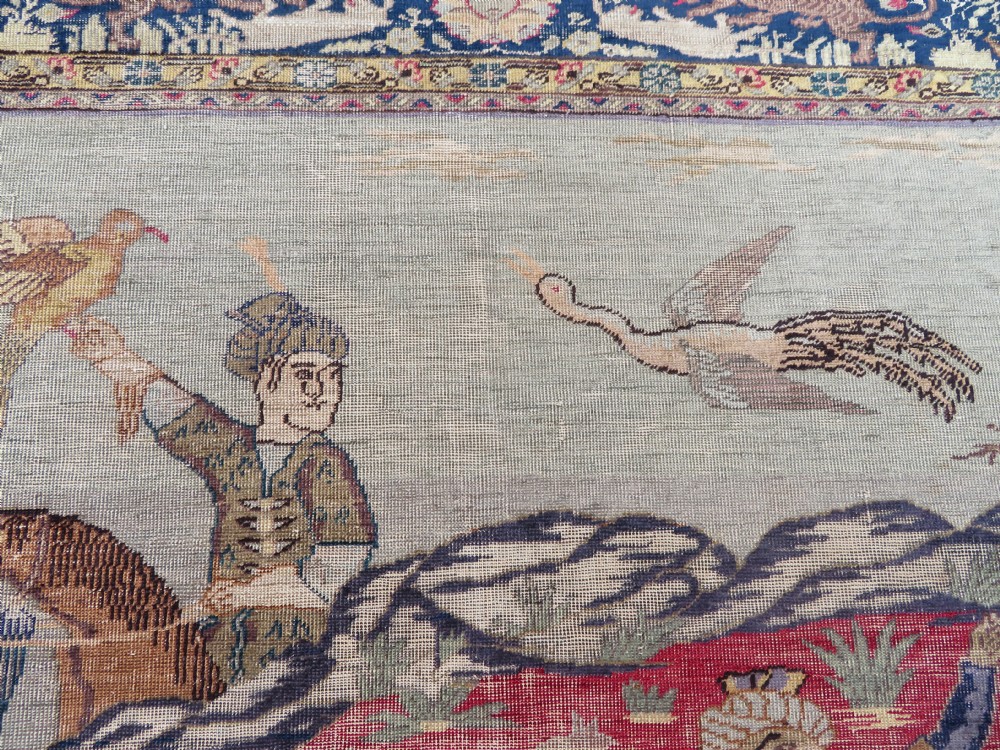A 19th-century West Anatolian silk rug featuring a red central panel depicting horseback hunters, surrounded by a floral and animal motif navy border. View V