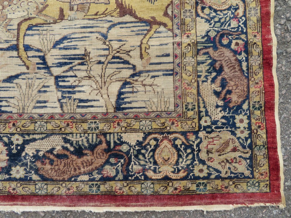 A 19th-century West Anatolian silk rug featuring a red central panel depicting horseback hunters, surrounded by a floral and animal motif navy border. View X