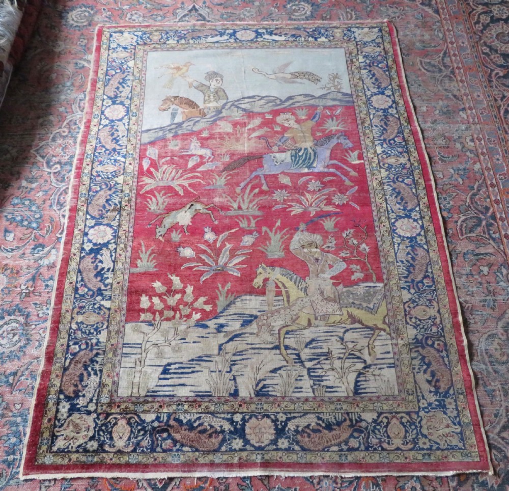 A 19th-century West Anatolian silk rug featuring a red central panel depicting horseback hunters, surrounded by a floral and animal motif navy border. View XVI