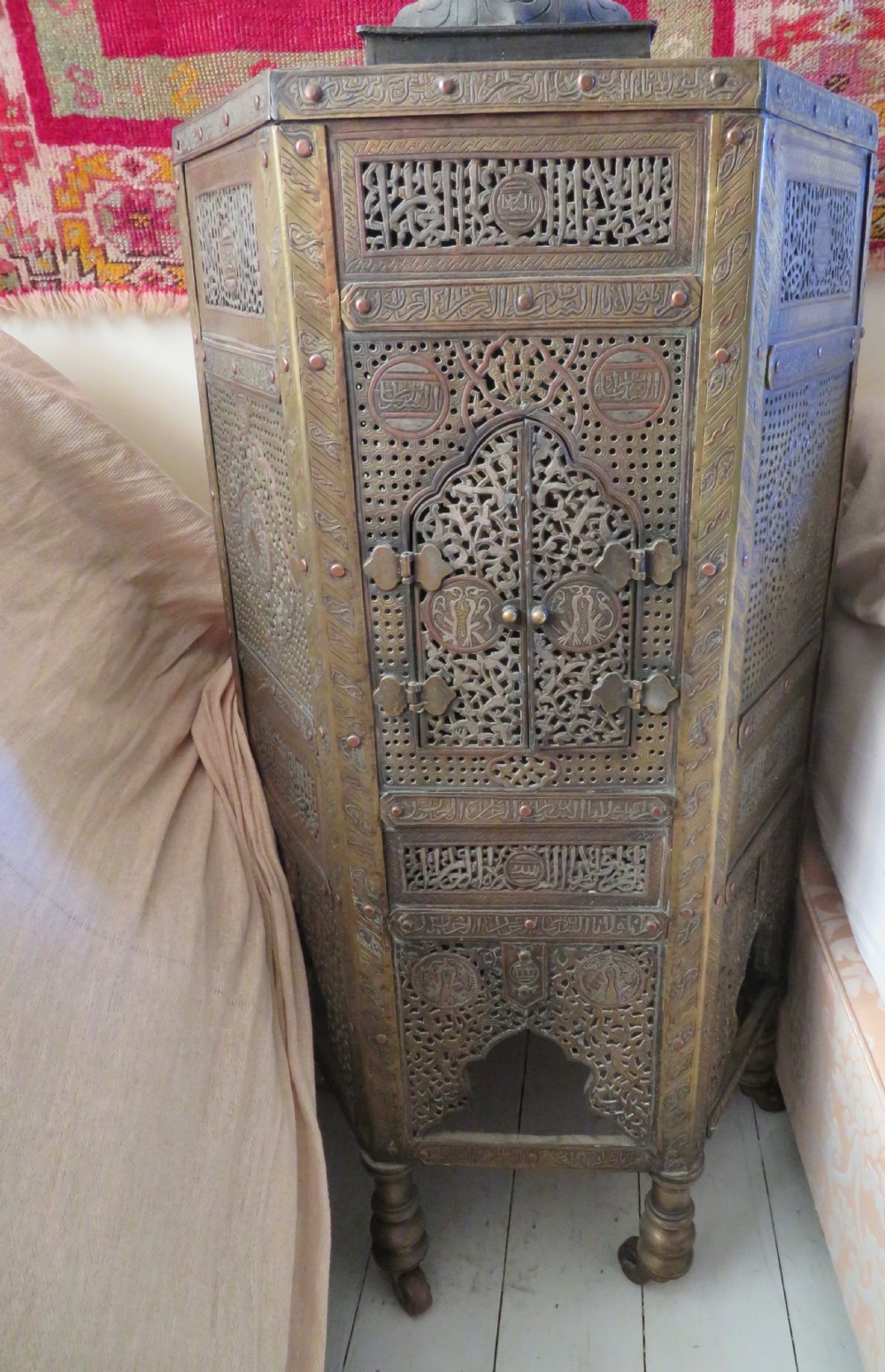A hexagonal antique brass occasional table with pierced openwork panels, Arabic calligraphic inlays in copper and white metal, and double cabinet doors.