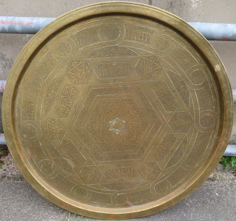 A large circular brass tray featuring detailed hand-engraved Islamic calligraphy, geometric interlace patterns, and a central star motif.