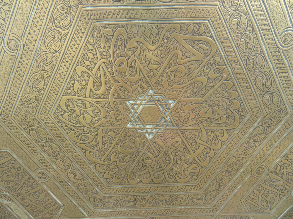 A large circular brass tray featuring detailed hand-engraved Islamic calligraphy, geometric interlace patterns, and a central star motif. View II