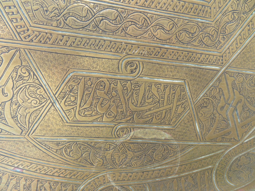 A large circular brass tray featuring detailed hand-engraved Islamic calligraphy, geometric interlace patterns, and a central star motif. View III