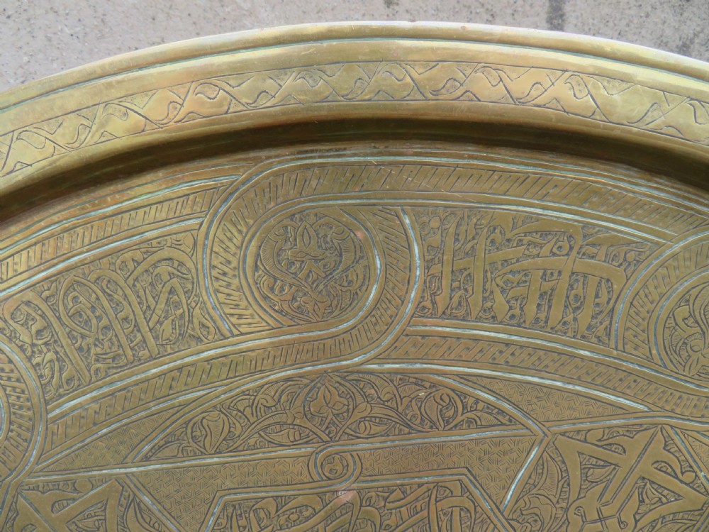 A large circular brass tray featuring detailed hand-engraved Islamic calligraphy, geometric interlace patterns, and a central star motif. View IV