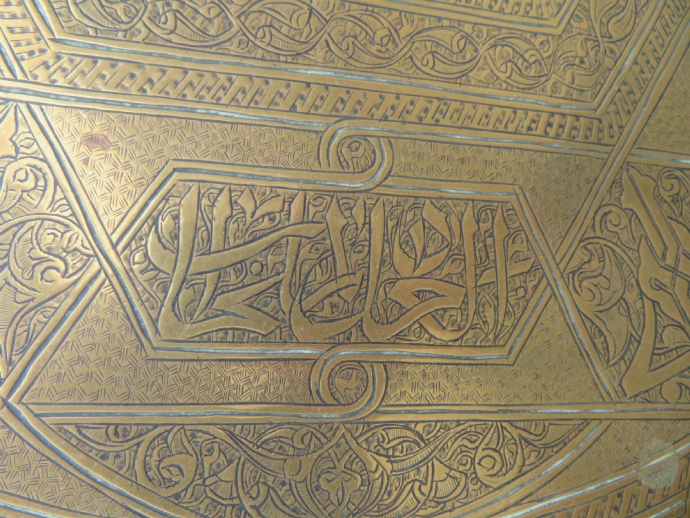 A large circular brass tray featuring detailed hand-engraved Islamic calligraphy, geometric interlace patterns, and a central star motif. View V