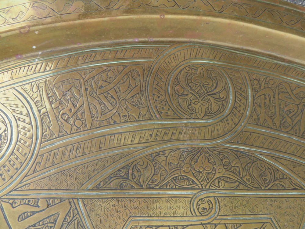 A large circular brass tray featuring detailed hand-engraved Islamic calligraphy, geometric interlace patterns, and a central star motif. View VI