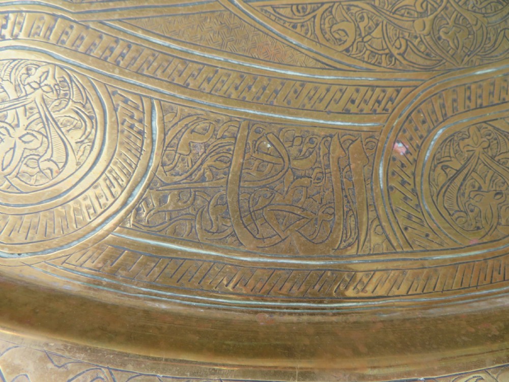 A large circular brass tray featuring detailed hand-engraved Islamic calligraphy, geometric interlace patterns, and a central star motif. View VII