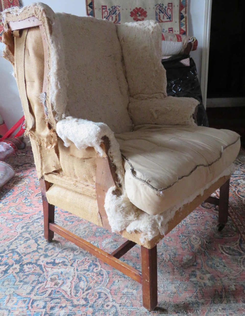 An antique Howard and Sons wingback armchair frame with exposed original stuffing, hessian backing, and square tapered wooden legs with castors.