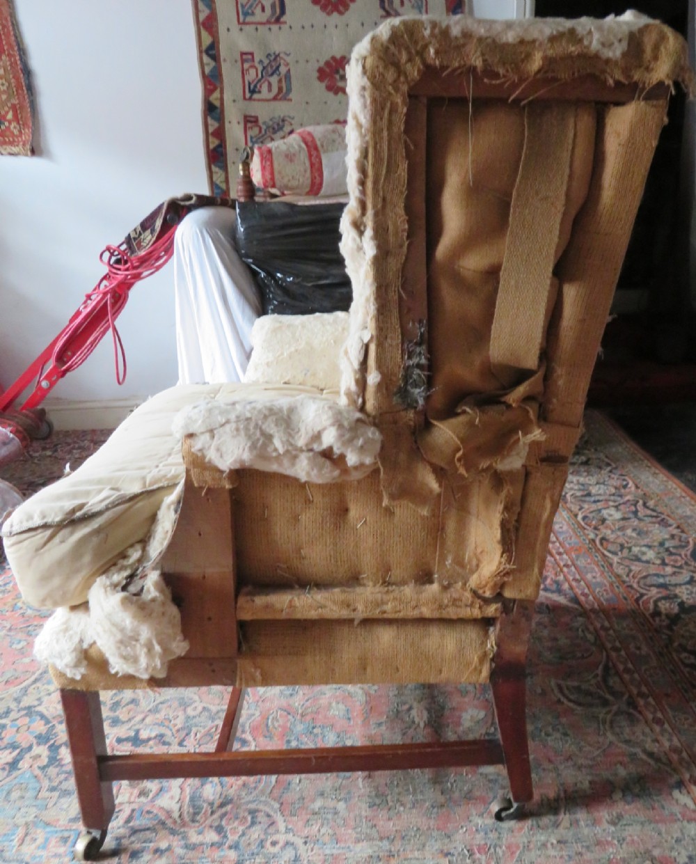 An antique Howard and Sons wingback armchair frame with exposed original stuffing, hessian backing, and square tapered wooden legs with castors. View II