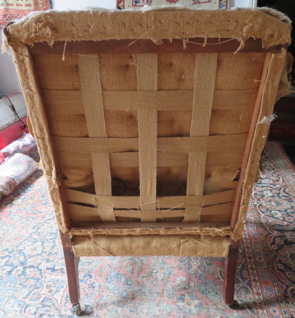 An antique Howard and Sons wingback armchair frame with exposed original stuffing, hessian backing, and square tapered wooden legs with castors. View III