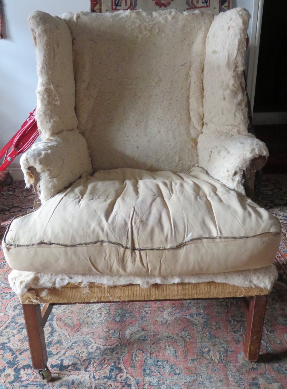 An antique Howard and Sons wingback armchair frame with exposed original stuffing, hessian backing, and square tapered wooden legs with castors. View IV