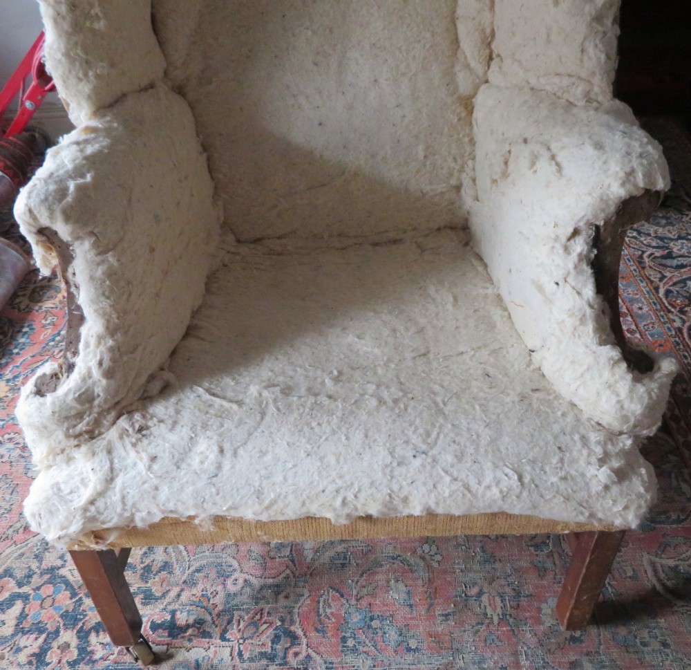 An antique Howard and Sons wingback armchair frame with exposed original stuffing, hessian backing, and square tapered wooden legs with castors. View V