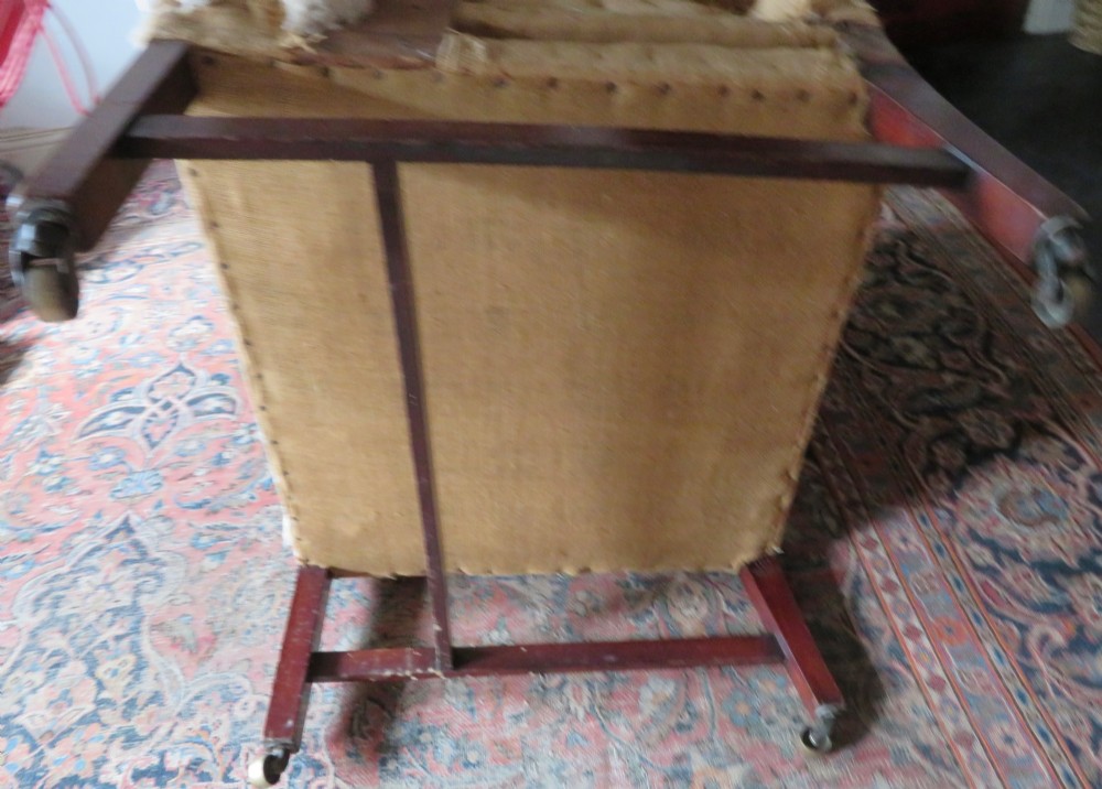 An antique Howard and Sons wingback armchair frame with exposed original stuffing, hessian backing, and square tapered wooden legs with castors. View VIII