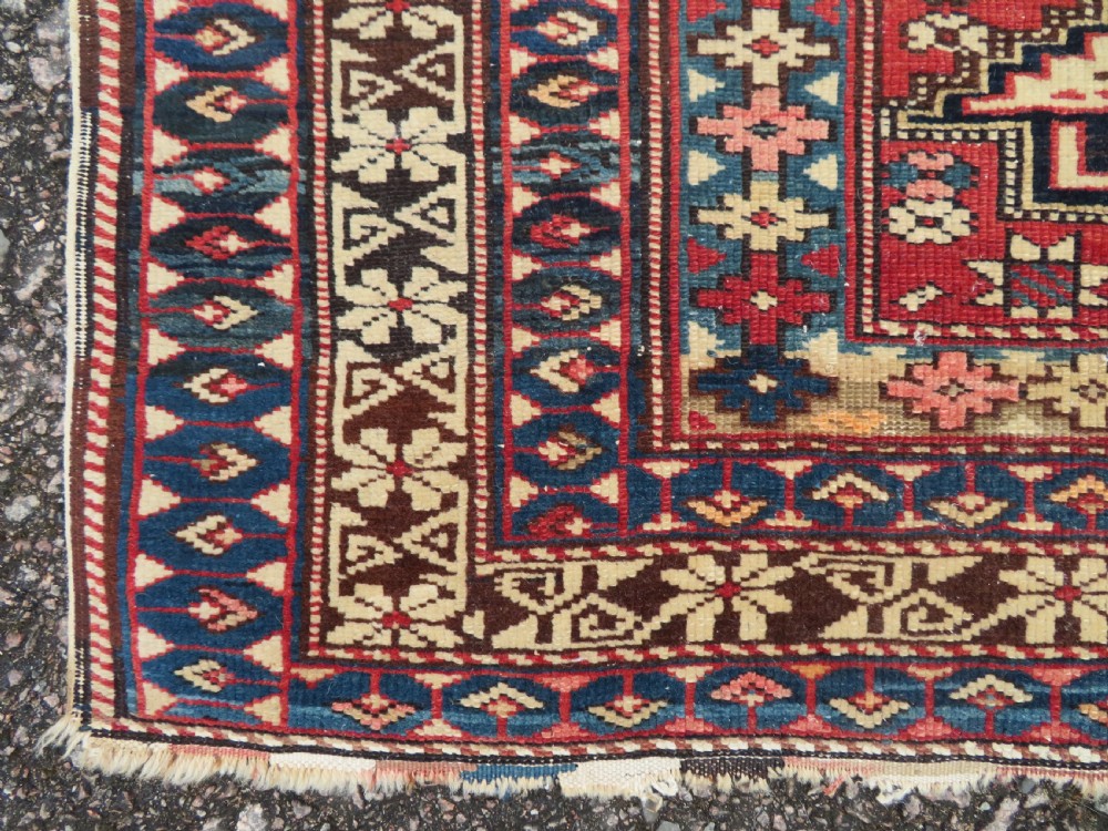 A distressed antique hand-knotted Caucasian Daghestan wool rug featuring geometric medallions and intricate borders in terracotta red, cream, and indigo blue. View V