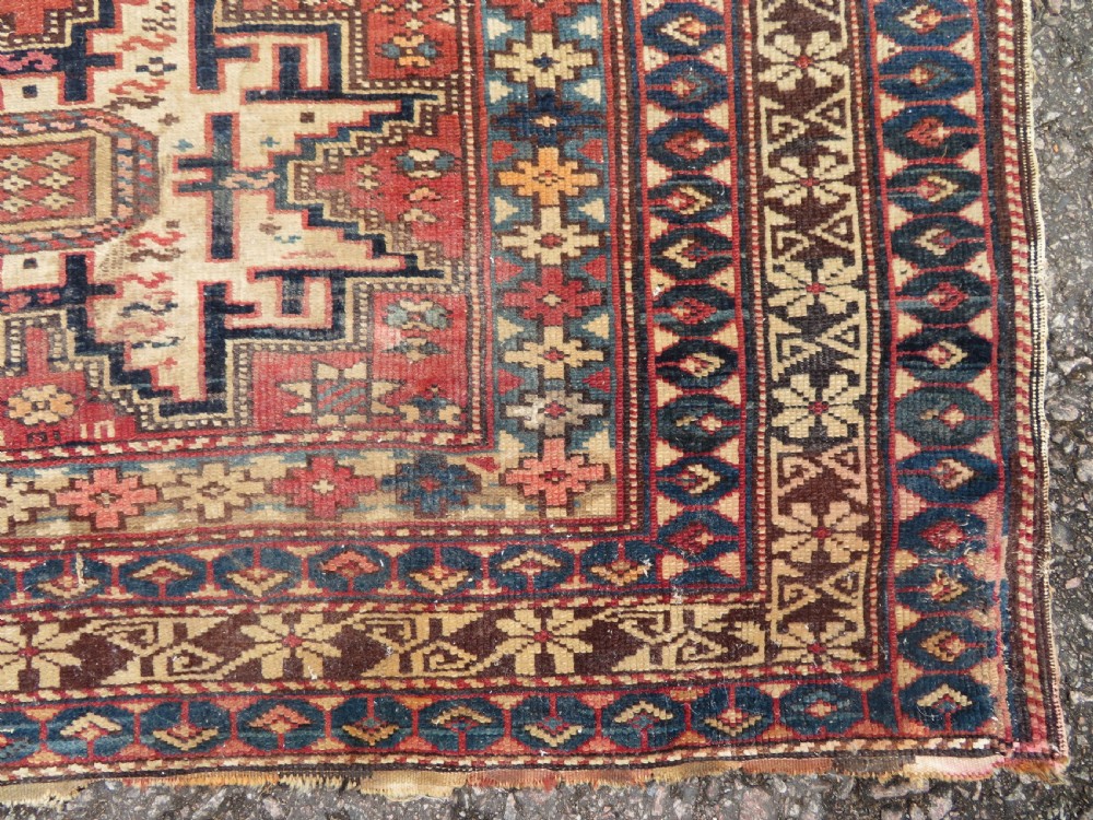 A distressed antique hand-knotted Caucasian Daghestan wool rug featuring geometric medallions and intricate borders in terracotta red, cream, and indigo blue. View VI
