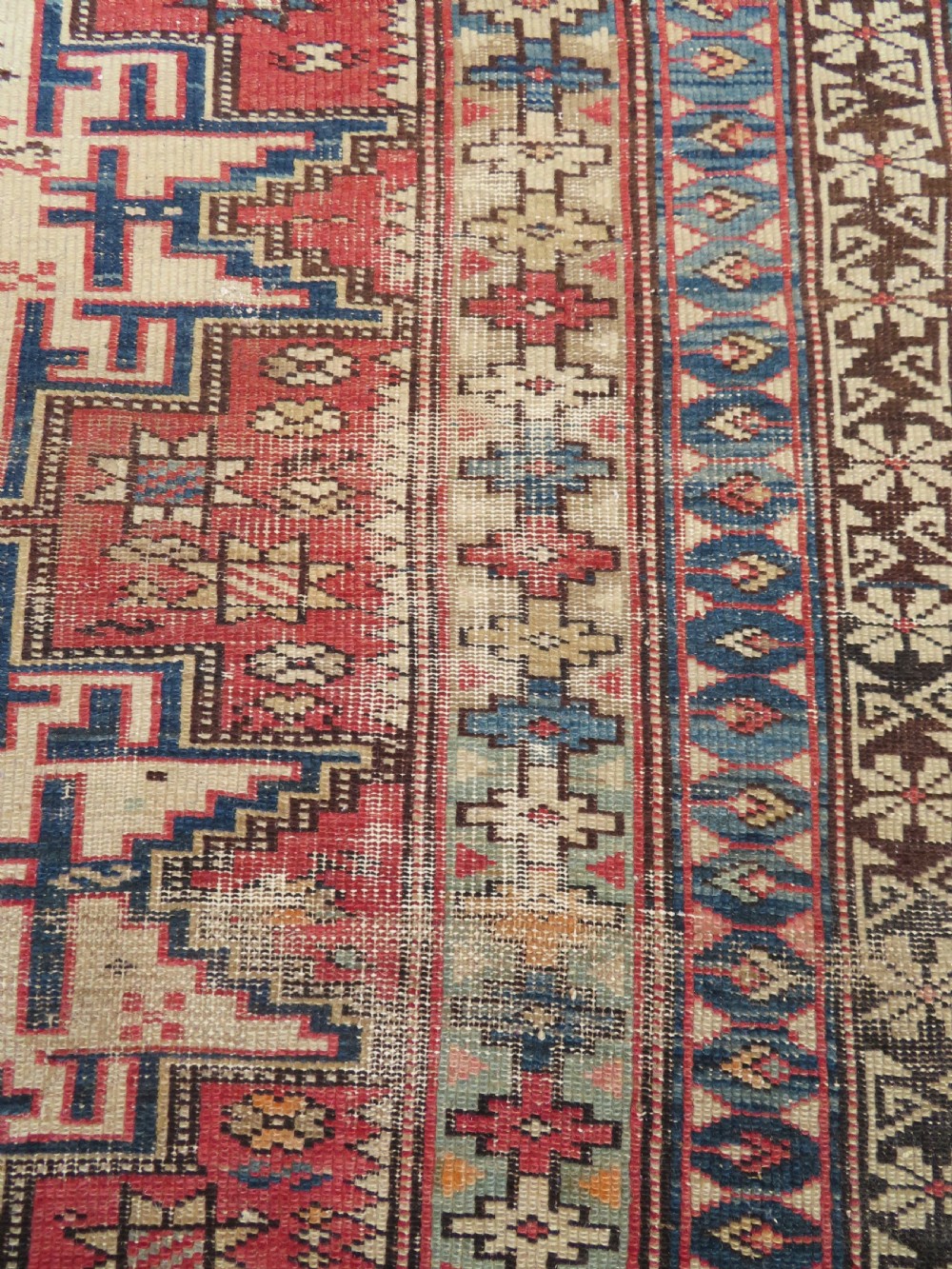 A distressed antique hand-knotted Caucasian Daghestan wool rug featuring geometric medallions and intricate borders in terracotta red, cream, and indigo blue. View VIII