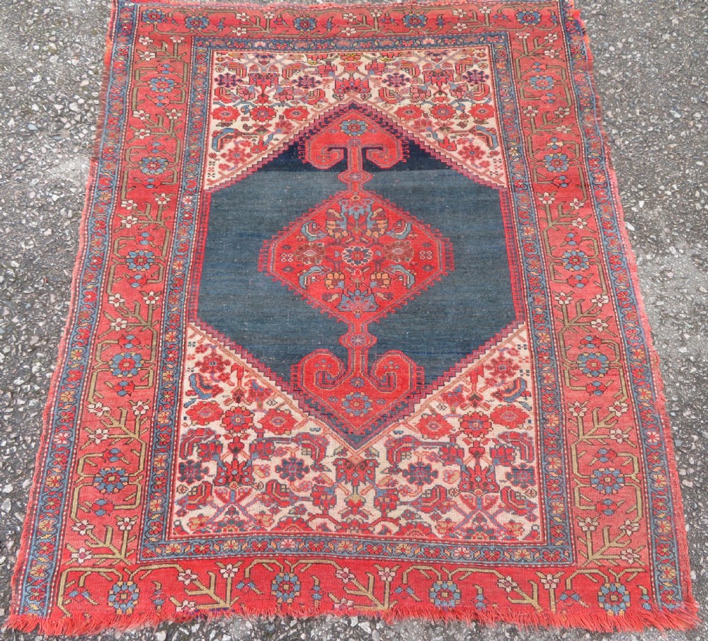 An antique hand-knotted Kurdish Bijar wool rug featuring a central red and blue medallion, cream patterned spandrels, and a terracotta floral border.