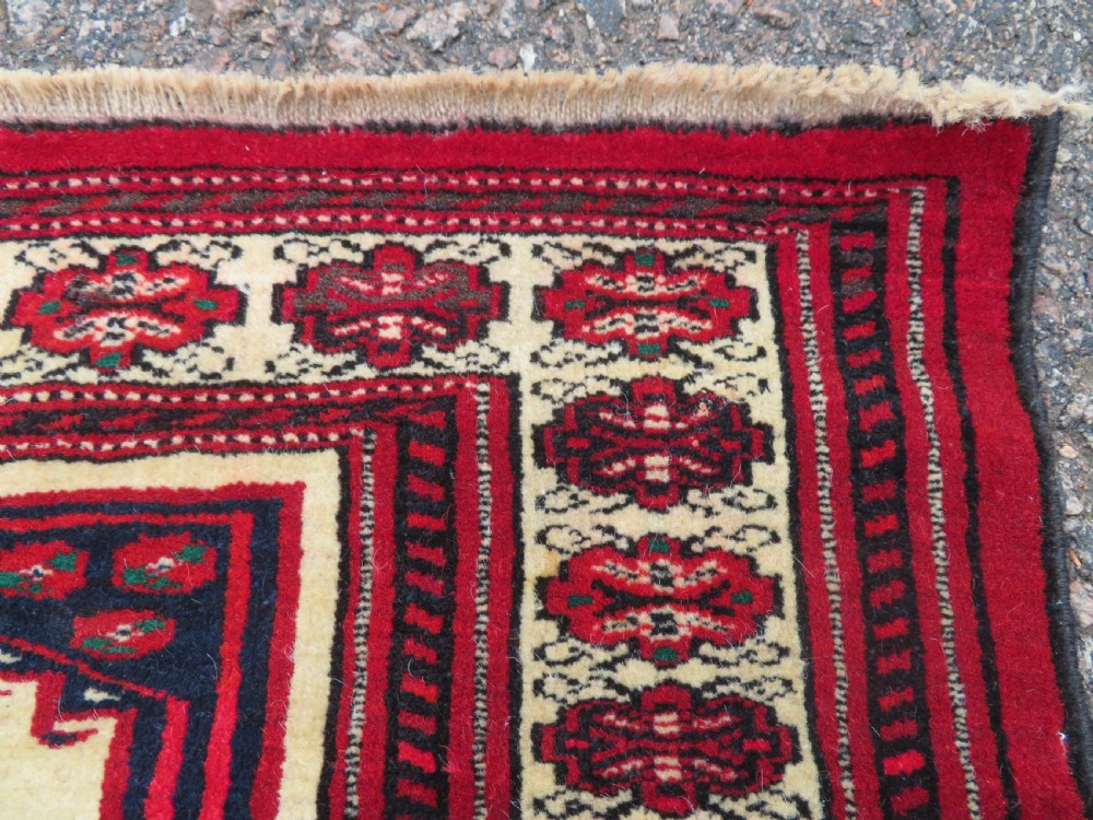 An antique rectangular hand-knotted Turkmen wool rug featuring red geometric diamond medallions on an ivory field with patterned dark borders. View VI