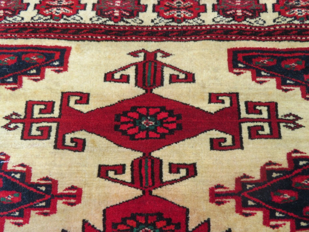 An antique rectangular hand-knotted Turkmen wool rug featuring red geometric diamond medallions on an ivory field with patterned dark borders. View IX
