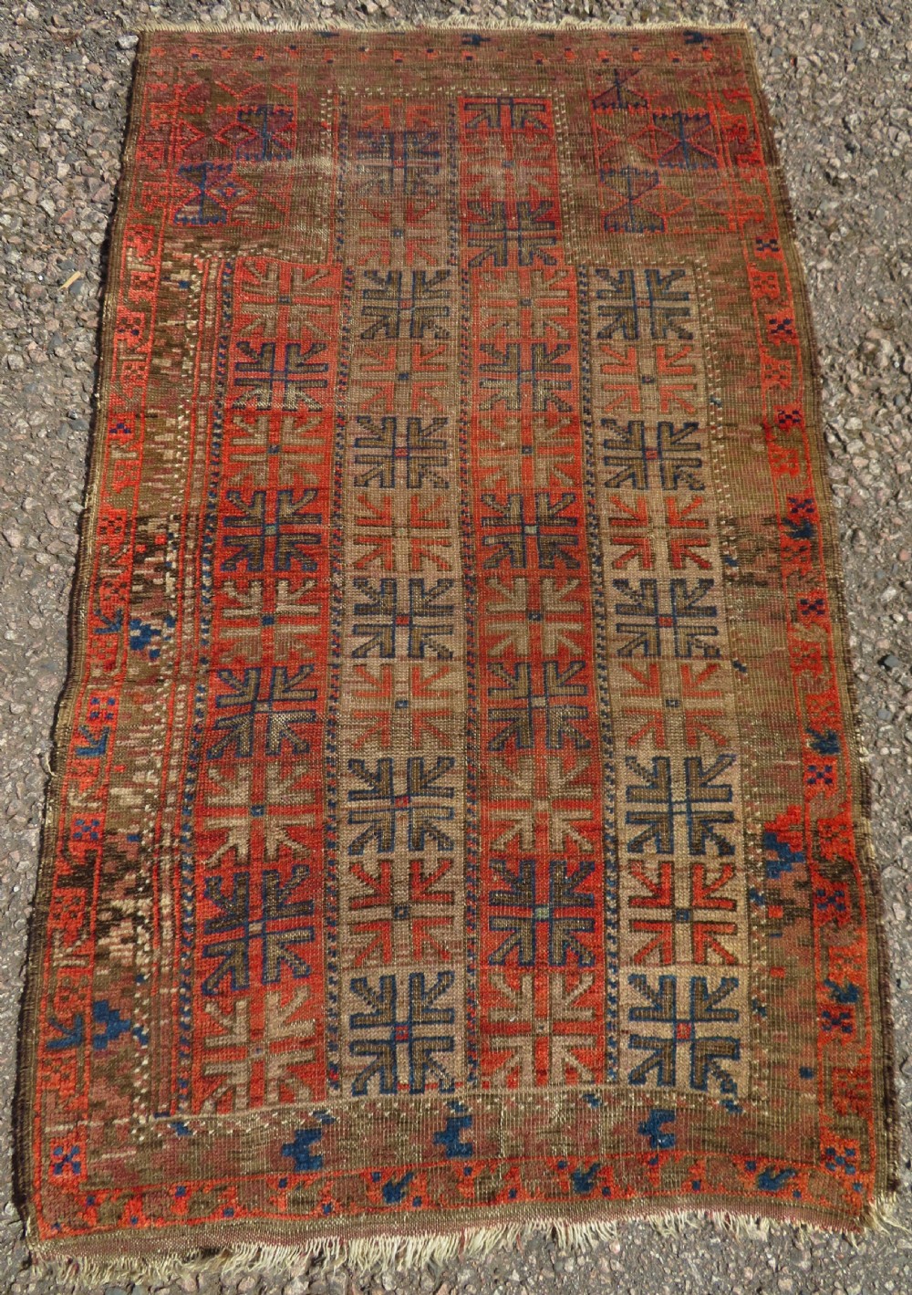 An antique hand-woven Baluch prayer rug showing distressed pile wear with geometric motifs in terracotta red, indigo, and deep brown.